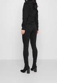 Mujer con jeans negros ajustados, botines negros de tacón y una camiseta negra de manga larga, de pie contra un fondo claro y liso.
