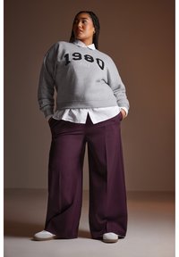 Grauer Sweatshirt mit "1980 PARIS" in Schwarz bedruckt, über einem weißen Hemd mit Kragen getragen. Dazu locker sitzende lila Hose und weiße Sneaker.