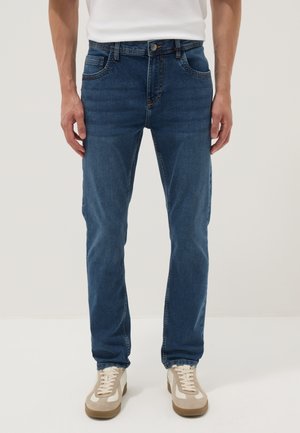 Hombre con jeans ajustados de mezclilla azul y zapatillas beige y blancas, de pie sobre un fondo claro y liso.
