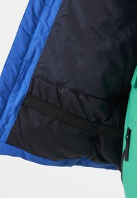 Izolowana kurtka z żywym niebieskim wykończeniem wewnętrznym, czarnym zewnętrzem oraz kieszenią na zamek błyskawiczny. Charakteryzuje się gładką teksturą i kontrastującymi kolorami.