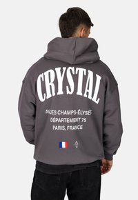 Felpa grigia con grande testo bianco che recita "CRYSTAL" e "AV. DES CHAMPS-ÉLYSÉS", con un dettaglio della bandiera francese e un logo a forma di diamante.