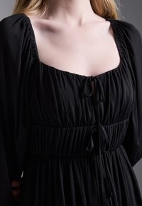 Robe noire avec un décolleté carré, tissu froncé et détail à cordon au niveau de la taille. De longues manches fluides mettent en valeur la texture douce.