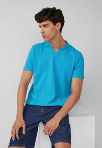 Turkusowa koszulka polo z krótkimi rękawami i nowoczesnym dekoltem w kształcie litery V, w połączeniu z ciemnoniebieskimi szortami dżinsowymi, na neutralnym tle.