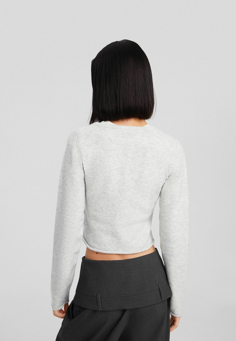 Grauer, kurzer Pullover mit langen Ärmeln, weichem Material und rundem Ausschnitt, kombiniert mit hochgeschnittenen, dunkelgrauen Hosen.