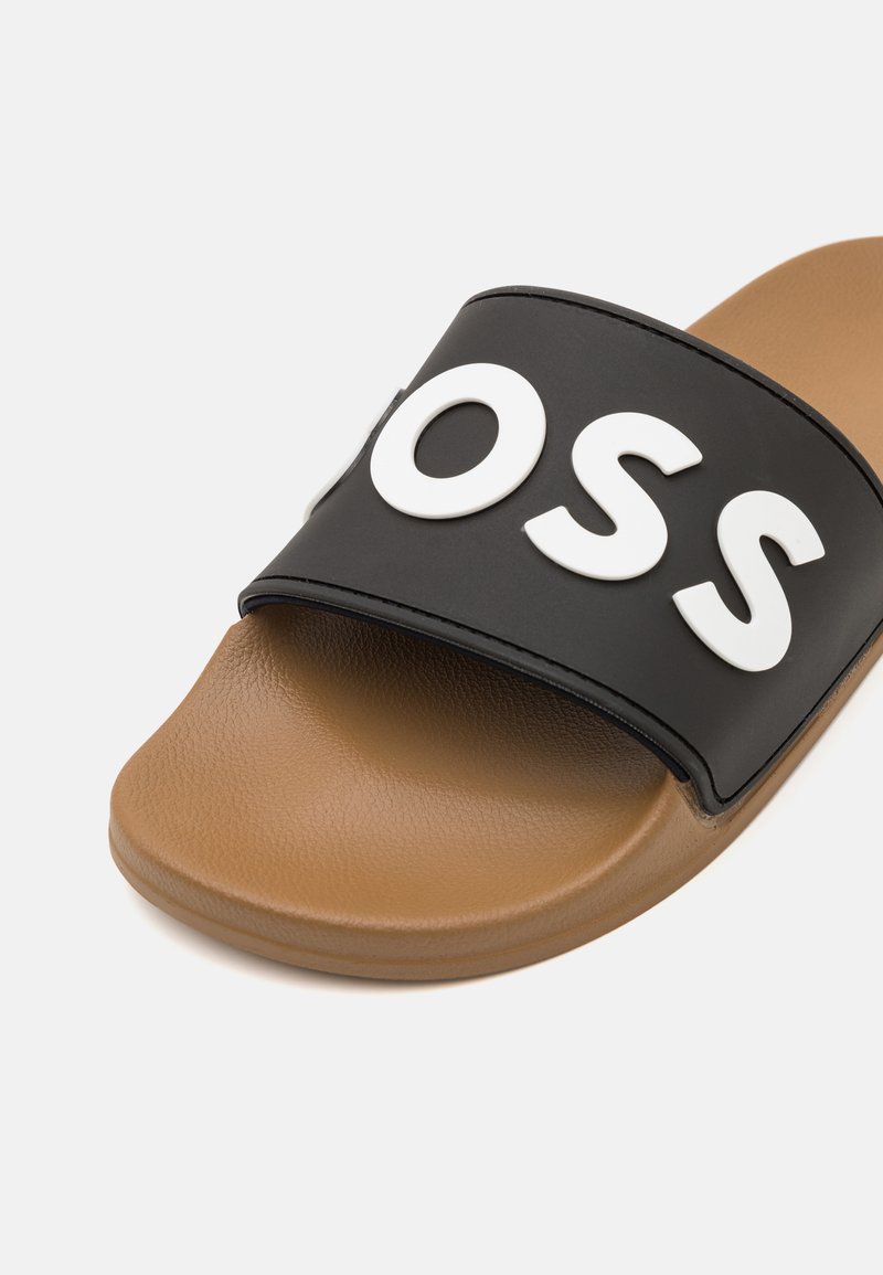 Sandale noire à glissière avec une large sangle arborant une lettre de couleur blanche audacieuse, reposant sur une semelle rembourrée couleur beige à texture lisse.