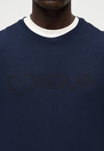 Άνδρας που φοράει μπλε σκούρο φούτερ με διακριτικό λογότυπο "DONDUP" και λευκό μπλουζάκι με στρογγυλή λαιμόκοψη ορατό στη γραμμή του λαιμού.