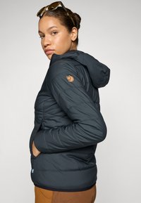 Doudoune à capuche bleu marine avec une texture matelassée et une patch logo sur l'épaule. Elle comprend des poches zippées et des poignets élastiques.