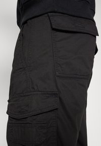 Närbild av svarta cargobyxor som visar ficklock med sömnadsdetaljer på lår- och höftområdet mot en neutral bakgrund.