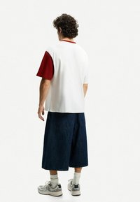 T-shirt bianco a maniche corte con un accento rosso su una spalla, abbinato a pantaloni corti in denim scuro larghi. Il modello è in piedi con le spalle rivolte all'osservatore.