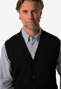 Schwarze gestrickte Weste mit V-Ausschnitt und fünf vorderen Knöpfen. Getragen über einem hellblauen und weißen gestreiften Hemd mit Knöpfen.