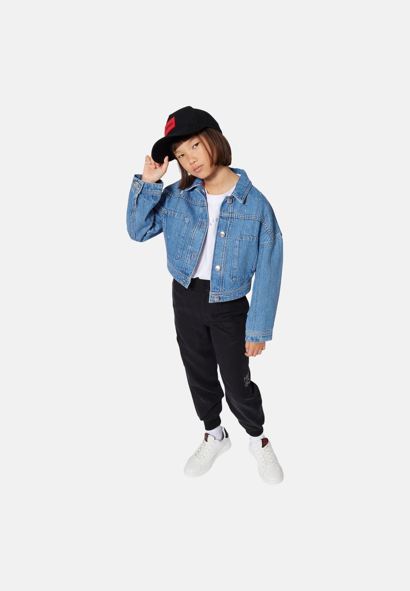 Lichtblauwe denimjack, cropped ontwerp met twee voorzakken, gecombineerd met zwarte joggingbroeken, witte sneakers en een zwarte pet met rode accenten.