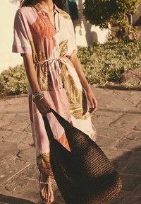 Kleid mit floralem Muster in knielanger Länge, mit Knopfleiste, kurzen Ärmeln und einem Bindegürtel. Kombiniert mit einer strukturierten, gewebten braunen Tragetasche.