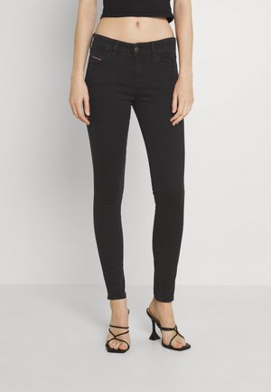 Kobieta w czarnych obcisłych dżinsach i czarnych sandałach na wysokim obcasie z paskami stoi na jednolitym białym tle.