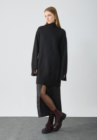 Grand pull noir à col montant, avec des poignets côtelés, associé à une jupe en cuir noire avec une fente sur le côté et des bottes bordeaux à lacets.