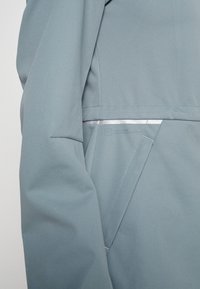 Manche de veste bleu clair et poche latérale avec une couture en angle et une bande réfléchissante étroite au-dessus de la poche.