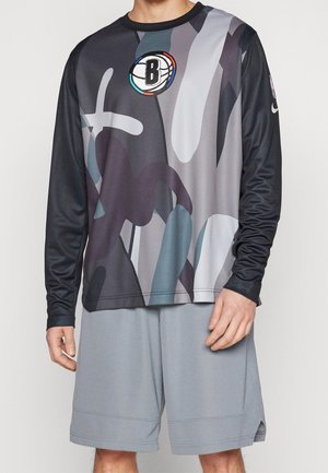 Mężczyzna w czarno-szarej, abstrakcyjnej koszulce koszykarskiej z długim rękawem i logo Brooklyn Nets oraz w jasnoszarych szortach.