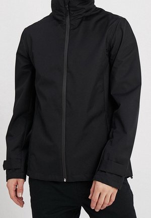 Person, der eine schwarze Jacke mit Reißverschluss, hohem Kragen und verstellbaren Manschetten trägt, vor einem einfarbigen hellen Hintergrund steht.