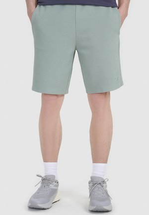 Mężczyzna ubrany w jasnozielone krótkie spodenki, szare sneakersy, białe skarpetki i ciemną koszulkę, stoi z rękami w kieszeniach na jednolitym tle.