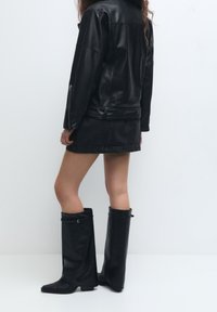 Persona vestita con giacca in pelle nera, mini gonna nera e stivali unici fino al ginocchio neri con flap con fibbie su uno sfondo bianco.