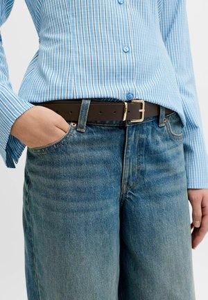 Persona con camisa de botones a rayas azules metida dentro de vaqueros azules con un cinturón marrón oscuro, mano izquierda en el bolsillo del vaquero.