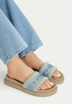 Piedi in jeans denim azzurri chiari che indossano sandali beige slip-on con cinturini blu con la scritta "G-STAR" su sfondo bianco.