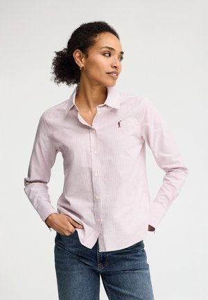 Woman with curly hair wearing a light pink striped button-up shirt and blue jeans, looking to her left with hands in pockets.