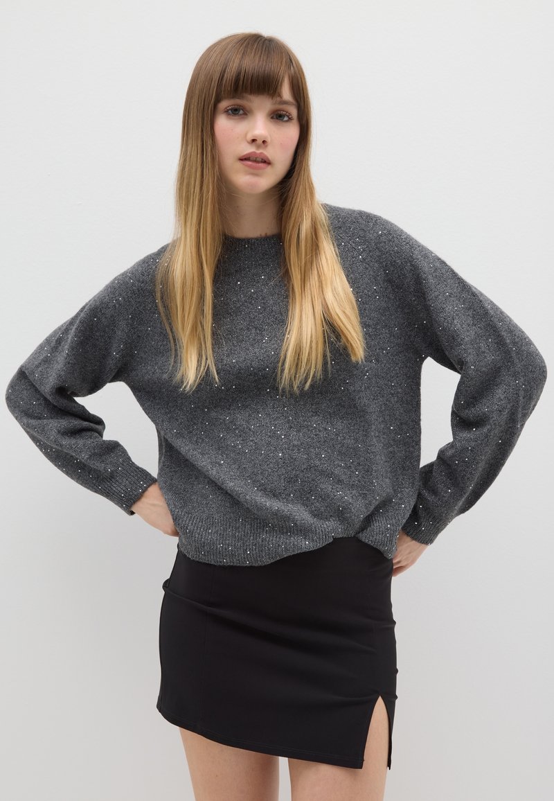 Pull en tricot gris avec des détails scintillants et des manches longues, associé à une mini-jupe noire avec une fente sur le côté.