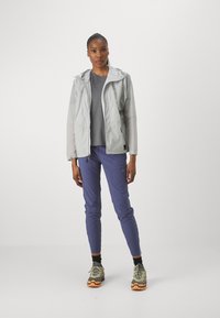Veste imperméable gris clair avec capuche, haut gris, pantalon violet et chaussures vertes avec semelles oranges ; texture lisse, design fonctionnel.