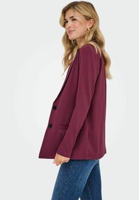 Burgundijas žakete ar piegriezumu, kurai ir divi melni pogas, viena kabata un gluda auduma. Valkāta ar ziliem džinsiem.