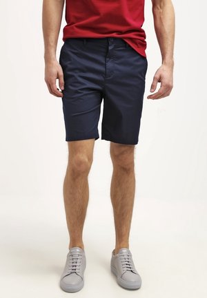 Mężczyzna w granatowych szortach, czerwonej koszulce i jasnoszarych sportowych butach, stojący na tle gładkiej białej ściany.