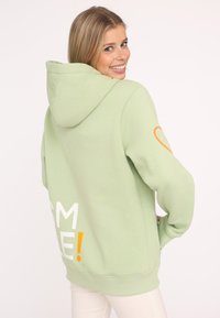 Mintgrüner Hoodie aus weichem Stoff, mit großen weißen Buchstaben "SMILE!" auf dem Rücken und einem orangefarbenen Herzakzent am Ärmel.