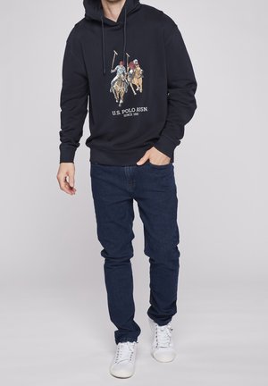 Marinblå huvtröja med ett framtryck av två polo-spelare på hästar. Kombinerad med mörka jeans och vita sneakers. Materialet verkar mjukt.