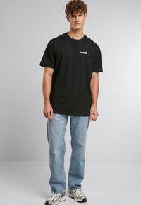 Schwarzes Baumwoll-T-Shirt mit kurzen Ärmeln, mit kleinem weißen "DEFSHOR"-Text auf der linken Brust, kombiniert mit hellblauen Jeans und Sneakers.