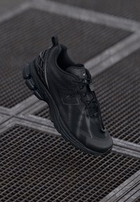 Chaussure de sport noire avec une tige en tissu maillé, des détails texturés, un système de laçage et une semelle rembourrée, arborant un logo réfléchissant sur le côté.