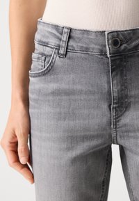 Grå denimjeans med en tettsittende design, som har en frontknapplukking, beltesløyfer og et svakt falmet mønster. Teksturert stoff er synlig.