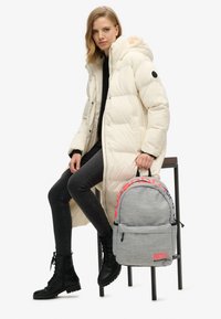 Mujer con un abrigo de plumas crema largo, jeans negros y botas sentada en un taburete sosteniendo una mochila gris de Superdry por la asa.