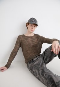 Brun, gennemsigtig strik bluse med lange ærmer og teksturerede striber, parret med grå camouflagedragt. En slidt grå kasket fuldender looket.