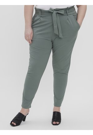 Woman wearing high-waist sage green tapered pants with fabric belt tie, paired with black open-toe heeled mules, standing with hands in pockets.