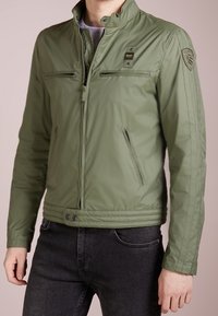 Veste légère vert olive avec un col montant, deux poches avant zippées et des rayures latérales discrètes. Finition du tissu texturée ; design ajusté.