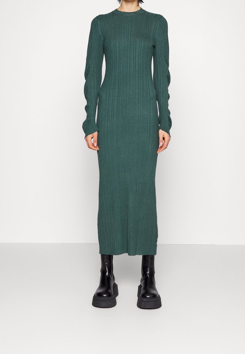 Personne portant une robe verte longue et moulante en maille côtelée avec des manches longues et des bottes noires épaisses à plateforme sur un fond blanc uni.