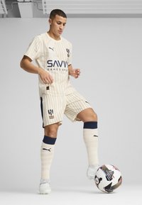 Equipación de fútbol a rayas beige con acentos azules, que presenta un logo y texto en la camiseta. Jugador con botas blancas, sosteniendo un balón de fútbol multicolor.