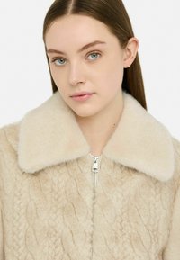 Cappotto di pelliccia beige con un ampio colletto morbido e un motivo a trecce testurizzato. Presenta una chiusura con zip e una leggera variazione tonale nel tessuto.
