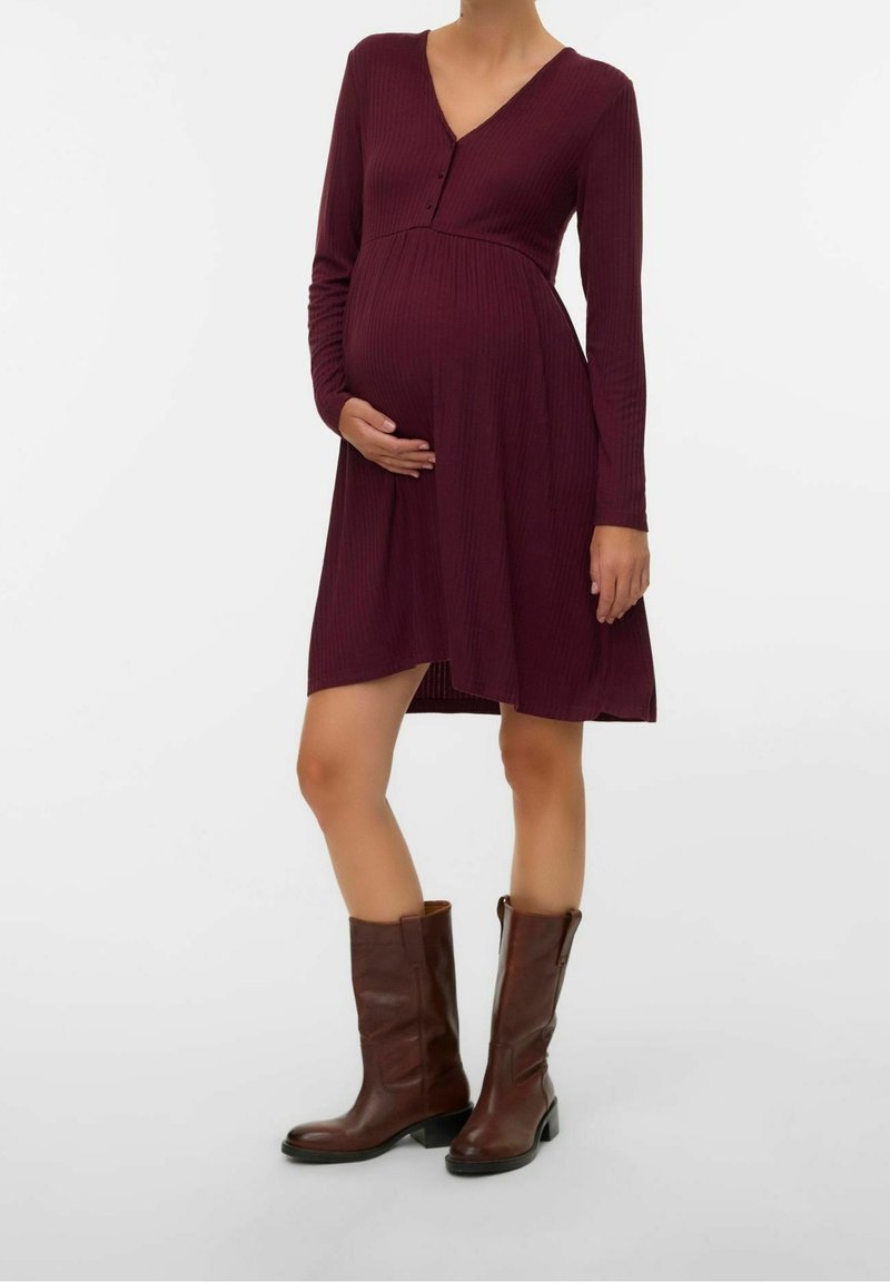 Femme enceinte portant une robe bordeaux à manches longues et des bottes marron mi-mollet, une main reposant sur son ventre.