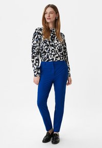 Bluzka z długim rękawem w czarno-biało-niebieski abstrakcyjny wzór, zestawiona z wąskimi, jednolitymi niebieskimi spodniami i błyszczącymi czarnymi loafersami.