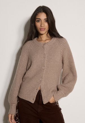 Young woman with long dark hair wears a taupe knitted button-up cardigan and dark pants, posing with one hand in pocket against plain background.