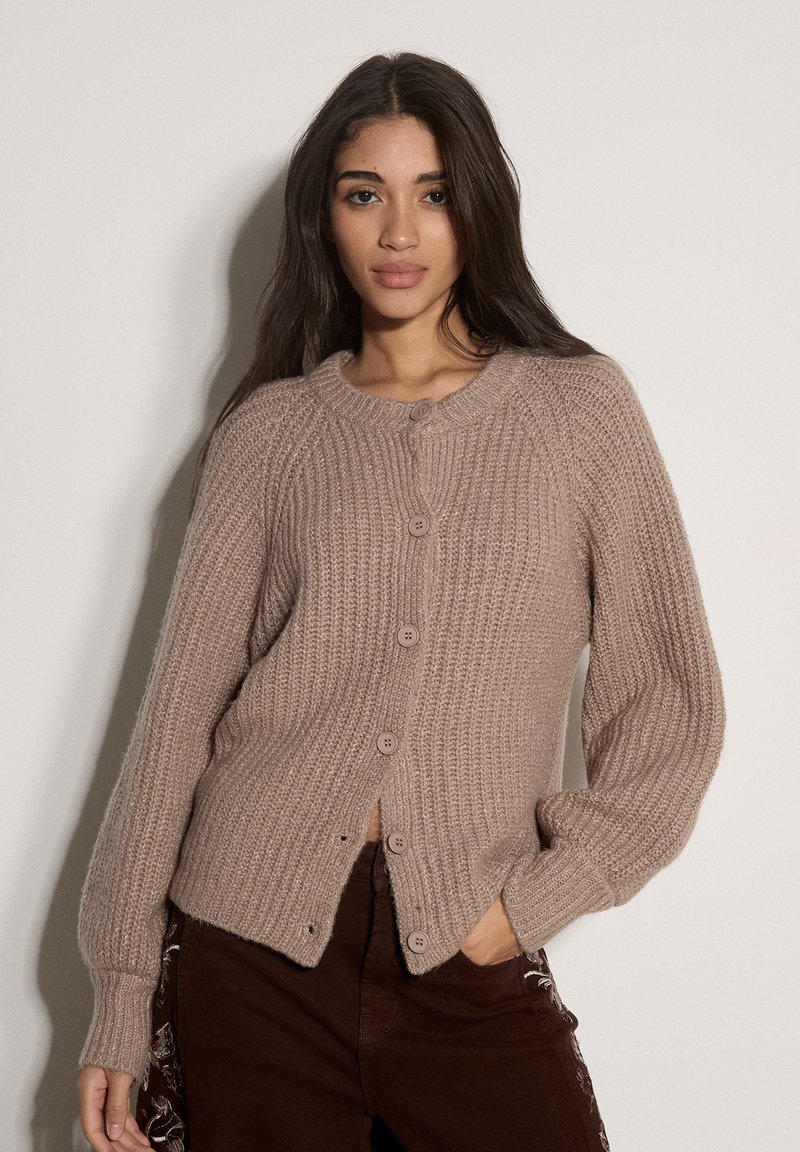 Jeune femme aux longs cheveux foncés porte un cardigan taupe en maille avec des boutons et un pantalon foncé, posant avec une main dans la poche sur un fond uni.