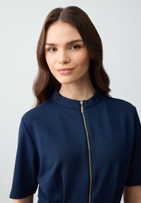Navyblauw kortmouwtop met een staande kraag en een gouden rits aan de voorkant, gemaakt van een gladde stof met een lichte glans.