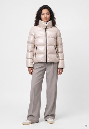 Frau mit dunklen Haaren, die eine glänzende beige Steppjacke, weite graue Hose und weiße Sneakers trägt, steht vor einem schlichten weißen Hintergrund.
