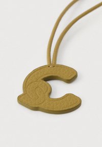 Pandantiv din piele texturată galbenă în formă de literă „C” stilizată, atașat de o sfoară asortată, pe un fundal alb.