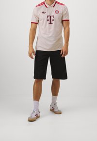 Mies, joka on pukeutunut beigeen Bayern Munich -jalkapallopaitaan, mustiin shortseihin, valkoisiin sukkiin ja valkoisiin Adidas-tennareihin, joissa on punaiset raidat, seisoo valkoista taustaa vasten.